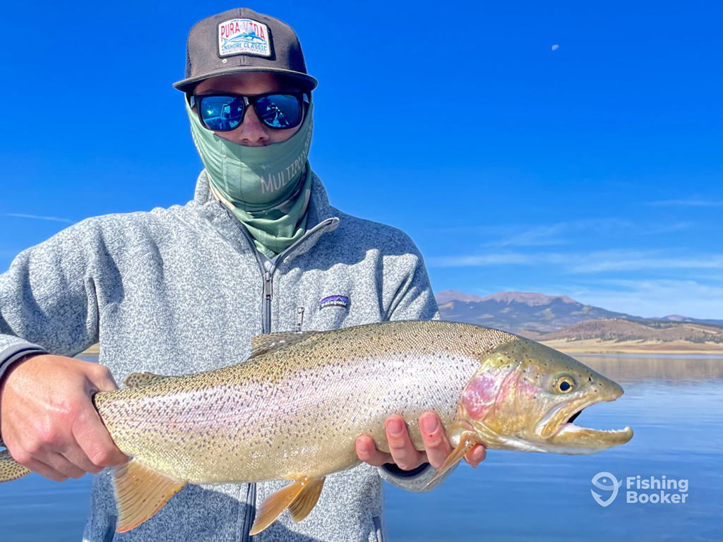 A closeup of a person’s holding a large Rainbow Trout on a lake with mountains in the background. They’re wearing sunglasses, a cap, and a neck gaiter.
