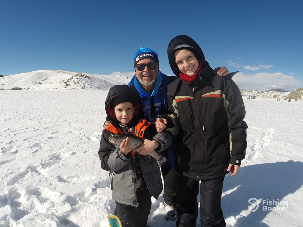 A man and two children are dressed in winter clothing, standing on a snowy landscape with clear skies. The child on the left is holding a small Trout.