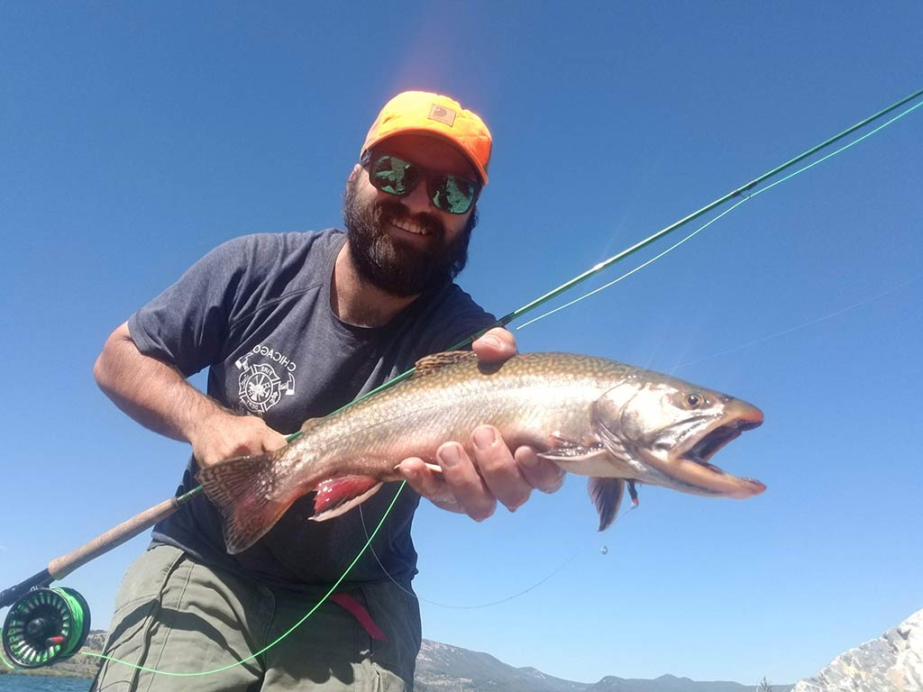 A man proudly holds a Brook Trout with his fly fishing rod, wearing an orange cap and sunglasses, against the clear blue sky.