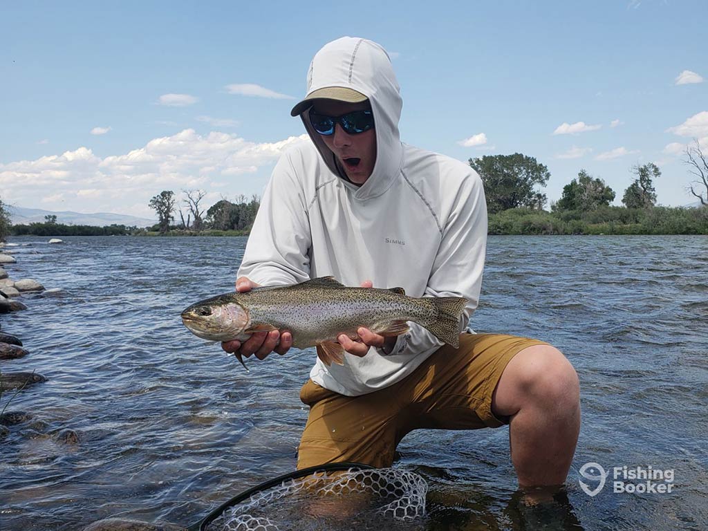 A surprised angler kneeling in the shallow waters of Montana, wearing a hoodie and sunglasses, while holding a sizeable Trout. Trees and a cloudy sky loom in the background.