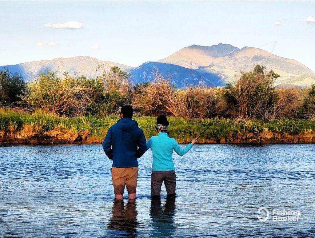 A view from behind of two, a man and a woman, standing in a river, facing away, casting their fly fishing lines into the waters of Montana. Majestic mountains and lush greenery are visible under a partly cloudy sky.