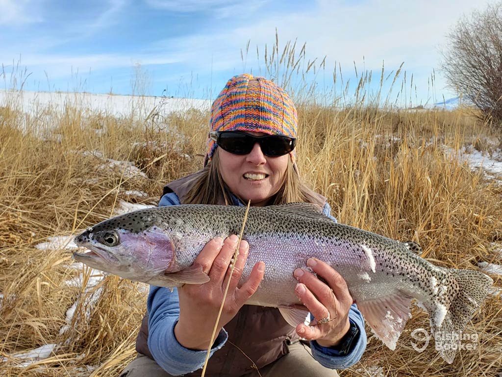 A woman in a bandana holding a large Trout, caught from a river or lake in Montana in winter, under a bright sky in a grassy field with snow patches visible.