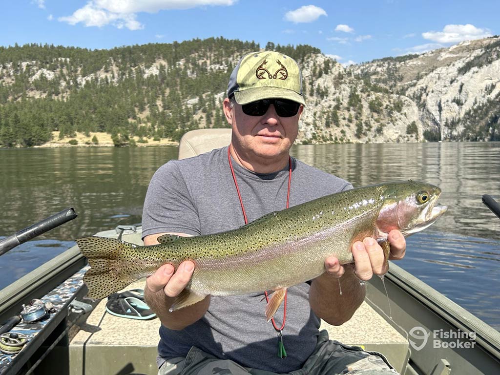 A person proudly holds a large Trout on a boat in a picturesque Montana lake, framed by forested mountains in the background.