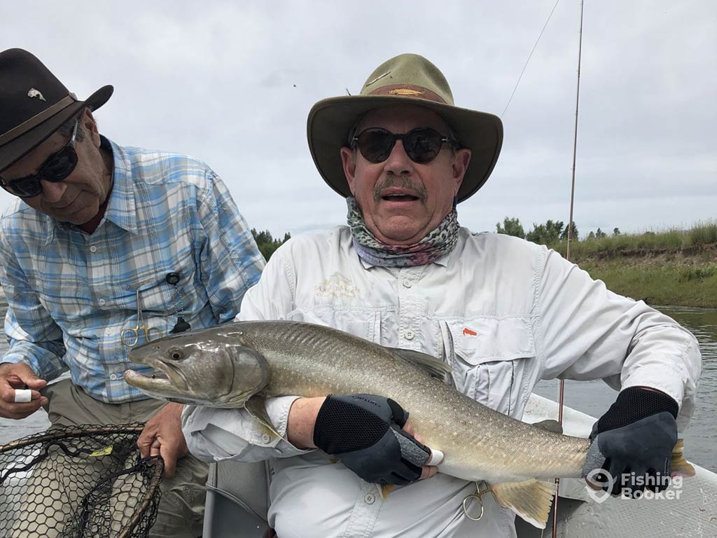 Two people are trout fishing in Montana on a boat. One is holding a large Bull Trout, and they're wearing sunglasses and a hat. The other is standing with a net against an overcast sky and grassy backdrop.