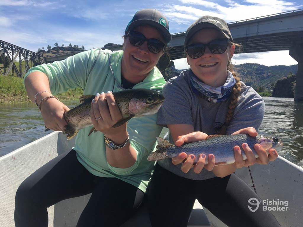 Two people are sitting in a boat, each holding a Trout, smiling against the picturesque backdrop of a Montana bridge and lush greenery.