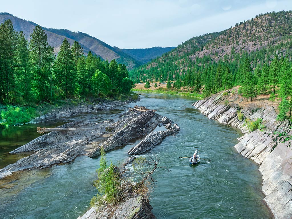 A lone kayaker is paddling in the distance along a river with rocky banks, surrounded by forested hills and distant mountains under a clear sky in Yellowstone National Park.