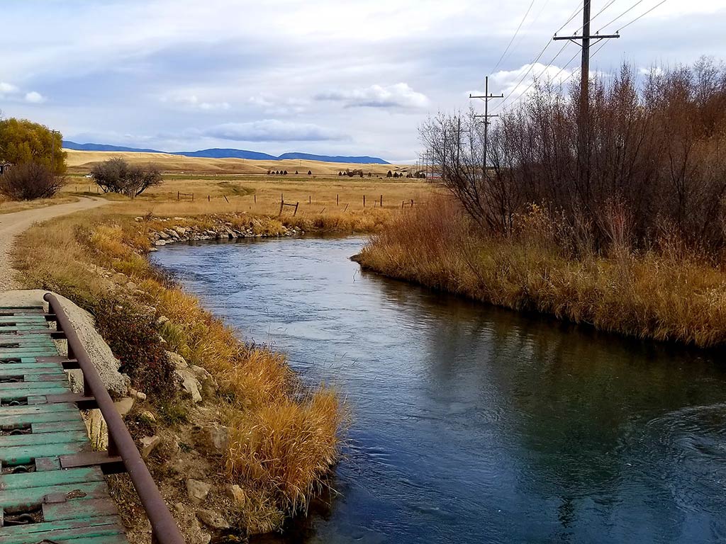 A narrow river near Lewistown, MT, flows alongside a grassy path, power lines, and a rural landscape under a cloudy sky.