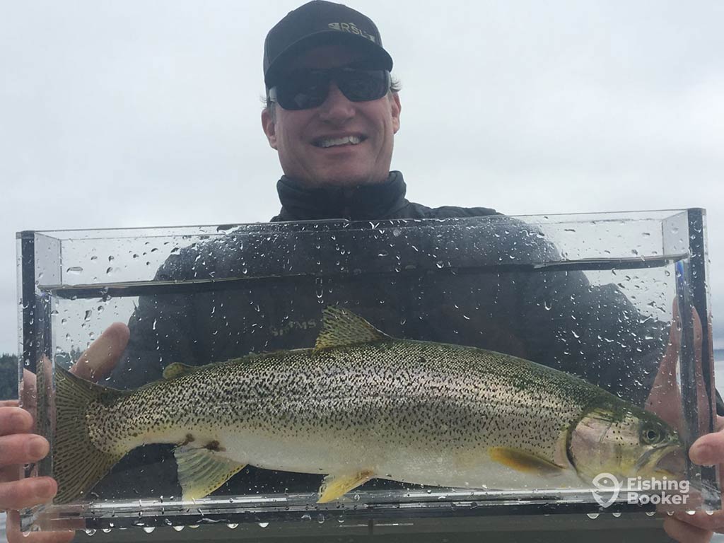 A person holding a large Trout encased in a clear container, wearing a dark jacket, sunglasses, and a cap. The fish has a mottled and speckled pattern.