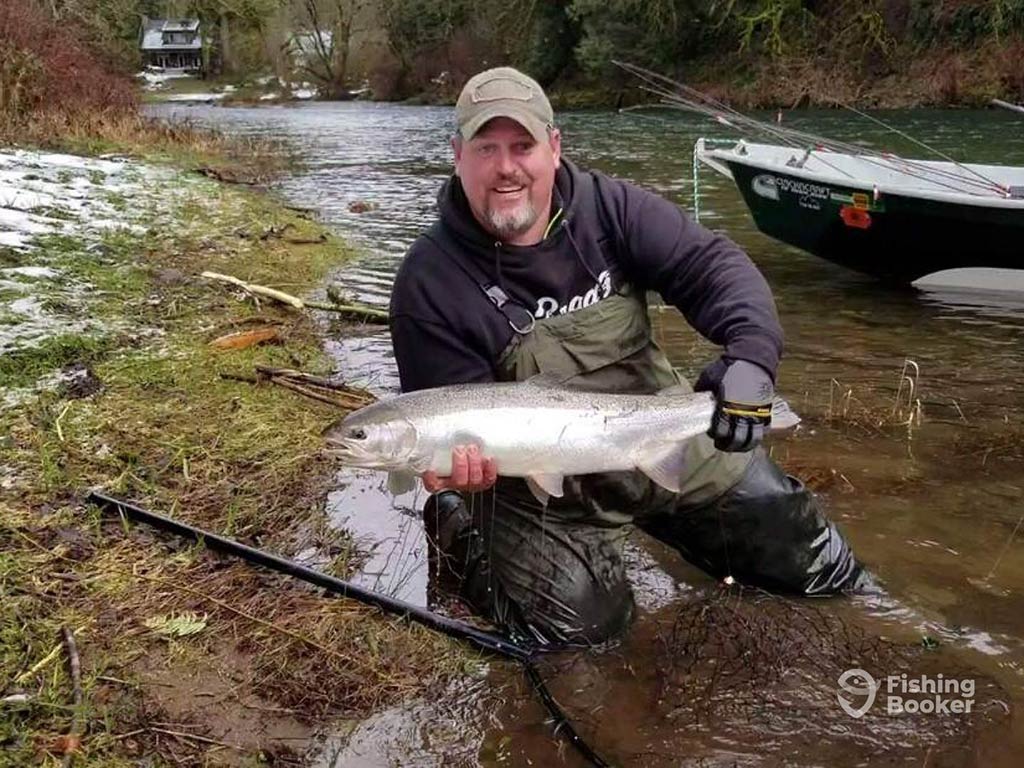 A person kneeling by a river in Washington, holding a large Steelhead fish. In the background, there’s a net and a boat.