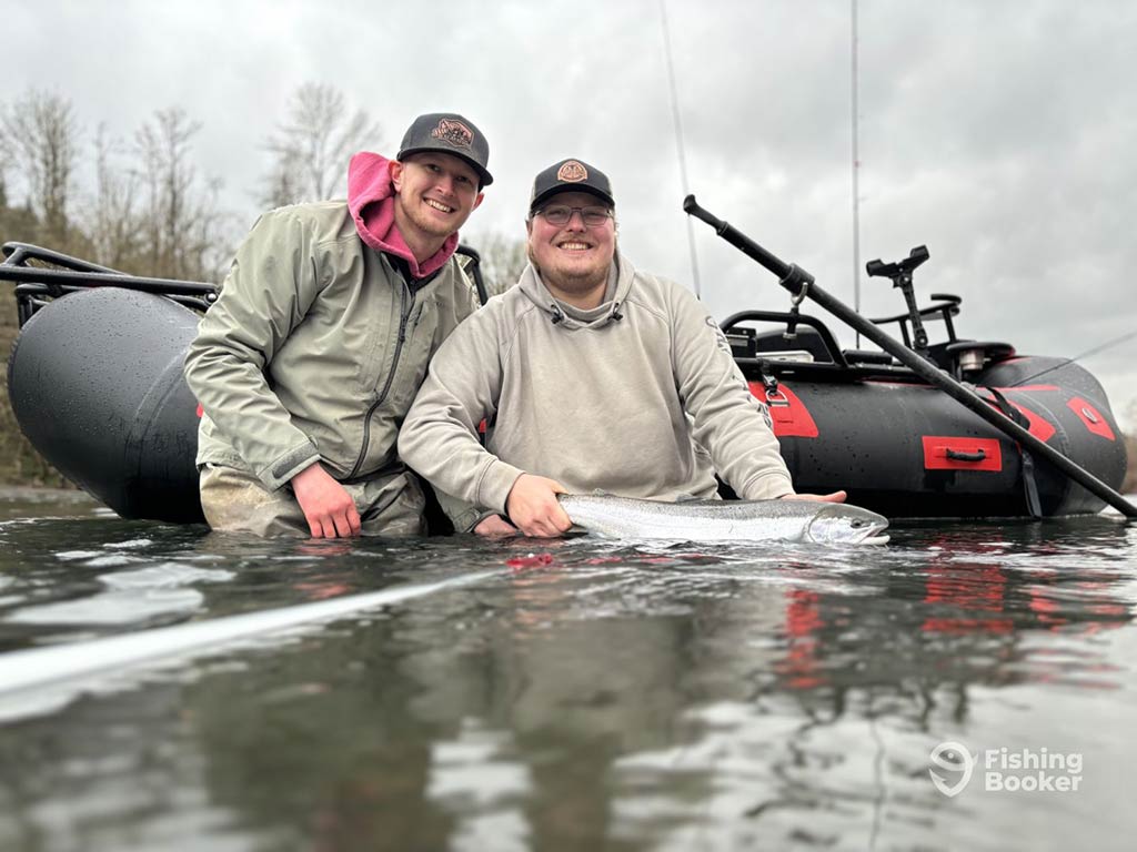 Two people in jackets are posing with a Steelhead in front of an inflatable boat on a cloudy day in a Washington river. You can see fishing rods visible on the boat.