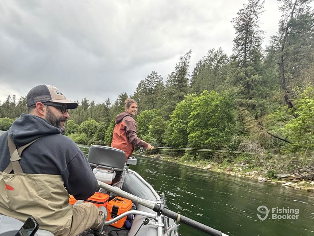 Two people are fishing from a boat on a river surrounded by trees, as they're hoping to catch some Trout. It's one of those perfect days where you don't need bright sunshine to enjoy the tranquility of nature. With any luck, they'll snag something worth telling stories about later!
