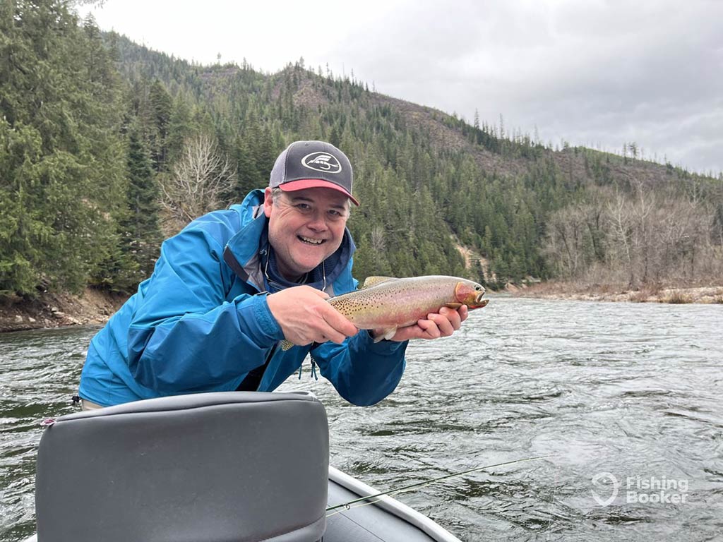 A man’s holding a Cutthroat Trout on a boat in a river in Washington, surrounded by forested hills on a cloudy day.