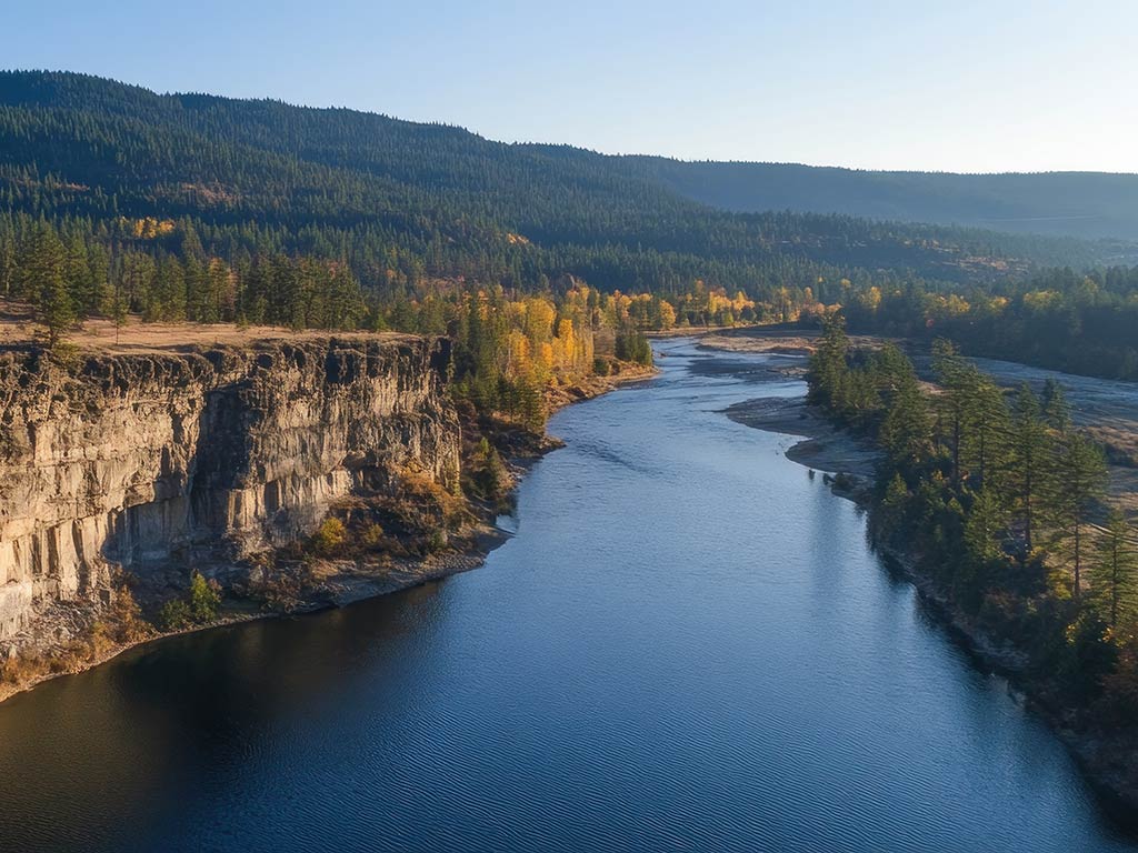 An aerial view shows the serene Kettle River in Washington, flanked by rocky cliffs and a lush forest with autumn foliage. Hills rise in the background under a clear sky.