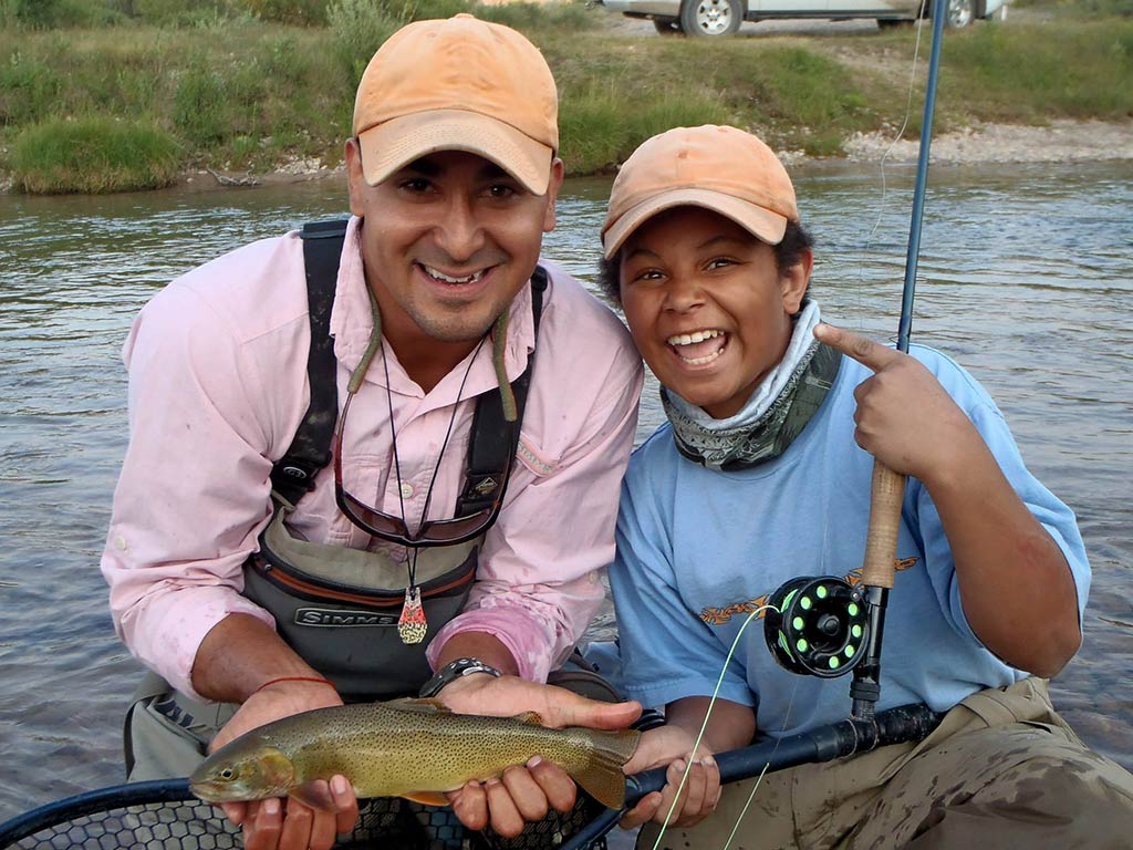 A man and a boy kneel by a river; one holds a Trout fish, while the other smiles and points at it. They’re both wearing caps and fishing gear, with a fishing rod in view.