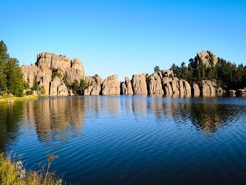 A clear blue lake reflects rocky cliffs and pine trees under a cloudless sky, offering classic Wyoming fishing experiences amid grass and wildflowers at the water’s edge.