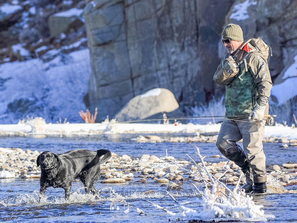 A man in waders and a beanie stands in a shallow, rocky river on a cold day, enjoying Wyoming fishing experiences as he watches his black dog playing in the water. He's hoping to spot Brown Trout or Rainbow Trout swimming by, knowing this stretch of river is famous for its Cutthroat Trout. The crisp air and peaceful surroundings make it easy to see why anglers come from miles around for the chance to land one of these prized fish.