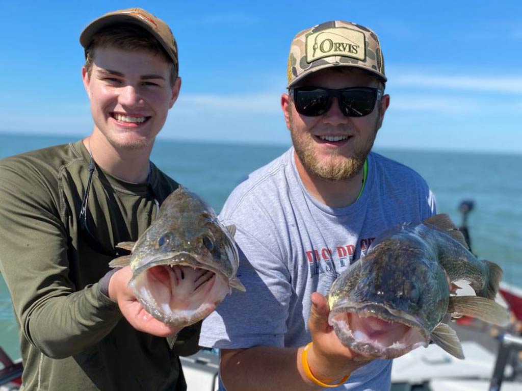 Two anglers are smiling and holding large fish each while enjoying Free Fishing Days on a charter boat with water and a blue sky in the background. Their excitement about reeling in these catches is obvious as they pose together for a photo during their memorable day on the water.
