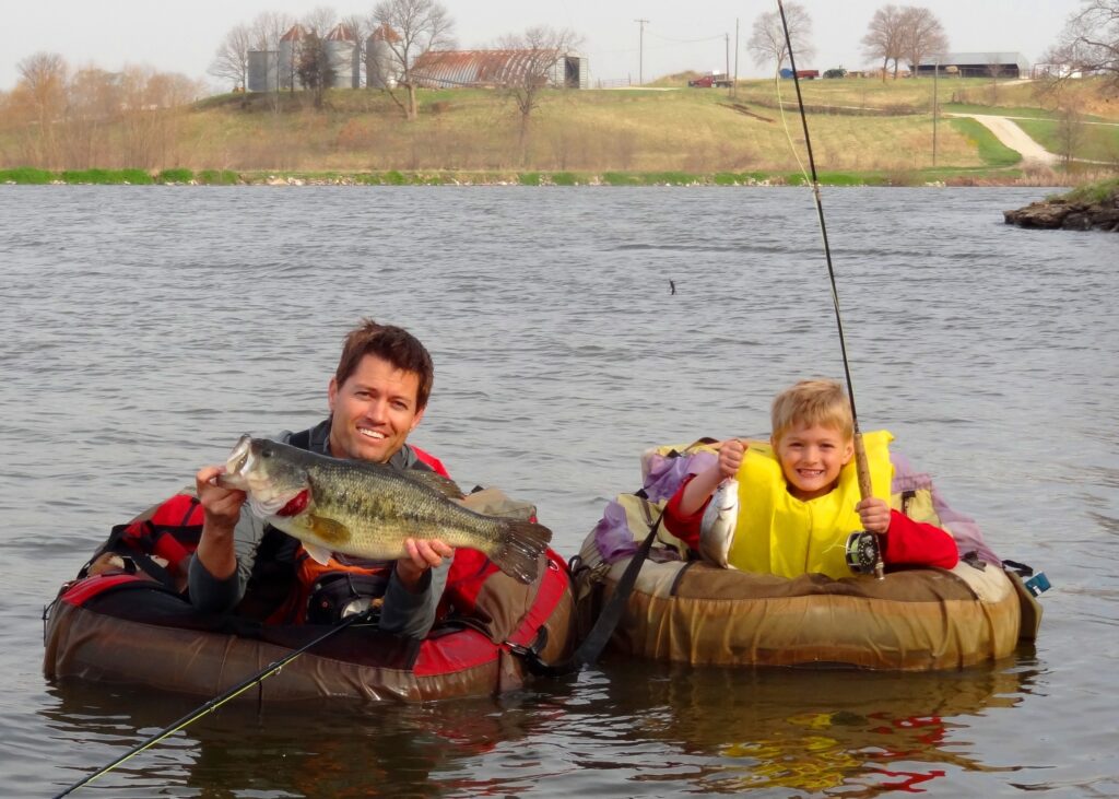 An adult and a child are float tube fishing on a lake, each holding a rod and their catch of a Largemouth Bass.
