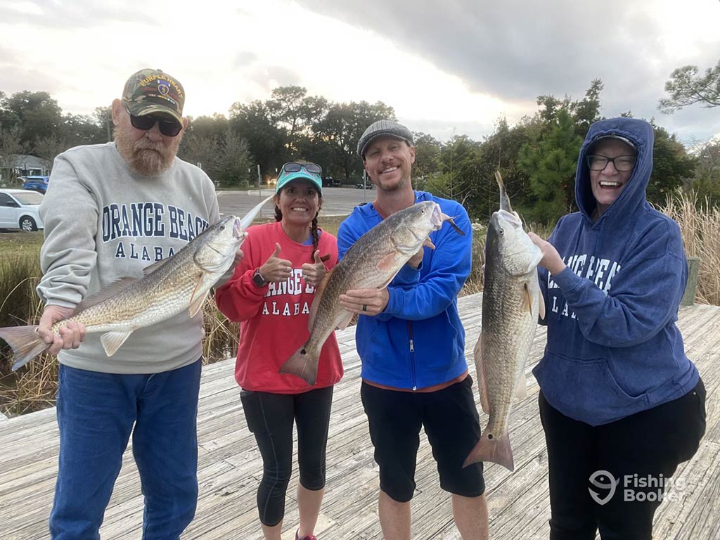 Four people are standing outdoors on a wooden dock, each holding a large fish and smiling at the camera—thanks to perfect Pensacola Fishing Tides. The background features trees and cloudy skies. The group proudly displays their catches, which include Red Snapper, King Mackerel, Grouper, and Amberjack.