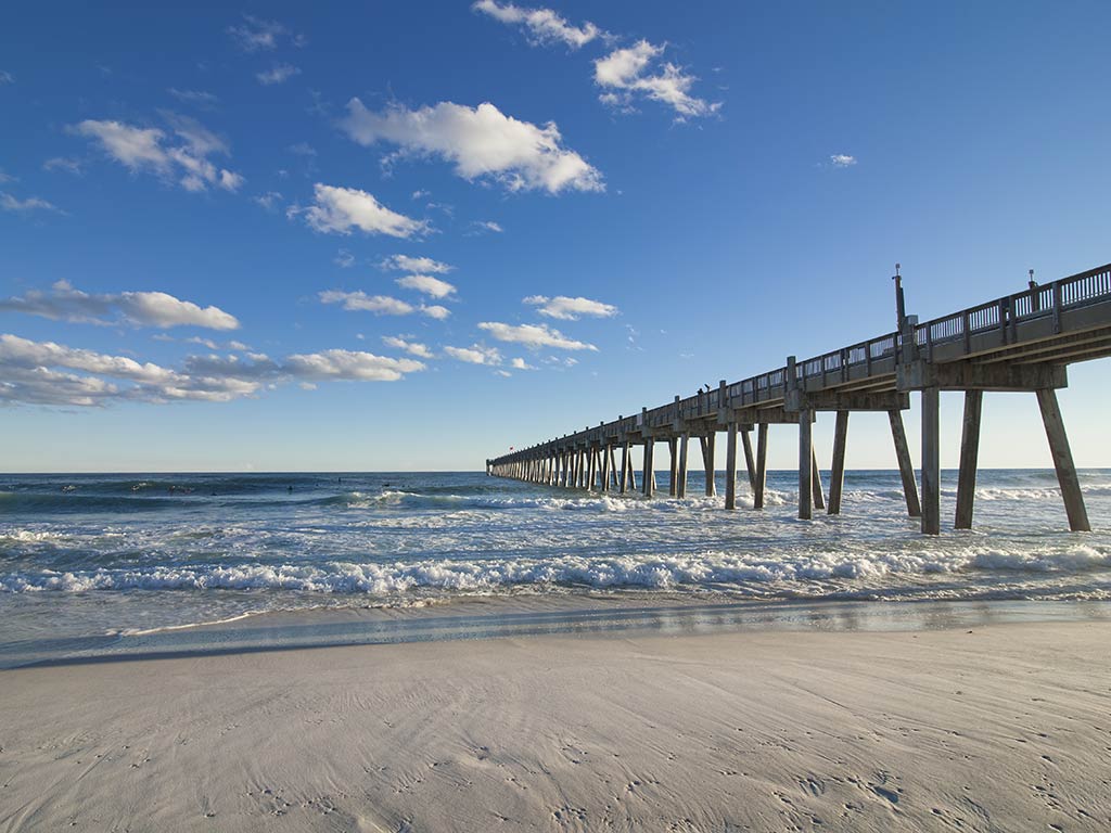 Pensacola's long pier stretches over the ocean beneath a blue sky with scattered clouds, while waves sculpted by the outgoing tide crash onto the sandy beach in the foreground.