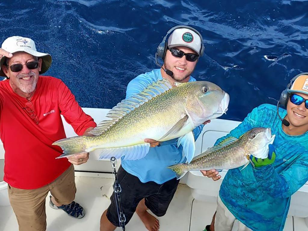 Three people on a boat, fishing for Tilefish, hold up large fish they’ve caught, smiling at the camera with the ocean in the background.