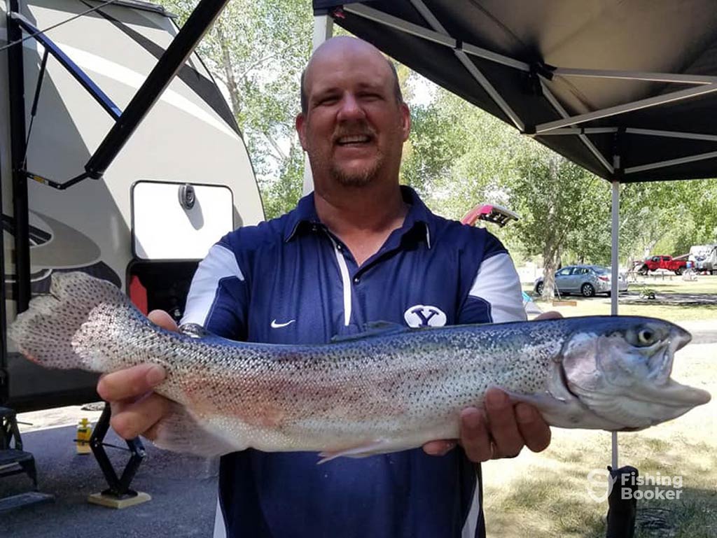 A man standing outdoors holding a large Rainbow Trout with both hands after trout fishing in Utah. There’s a trailer and some trees in the background.
