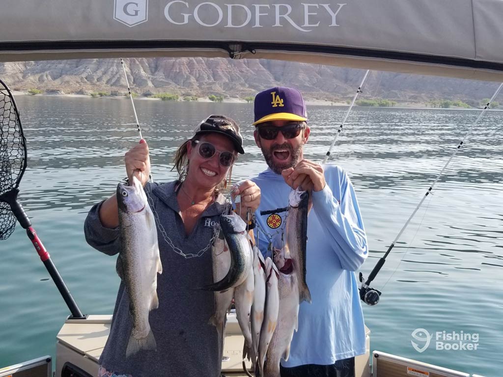 Two people are on a boat, each holding several caught Trout and smiling for the camera while Trout fishing in Utah. Behind them, a scenic lake stretches out with majestic mountains rising in the background.