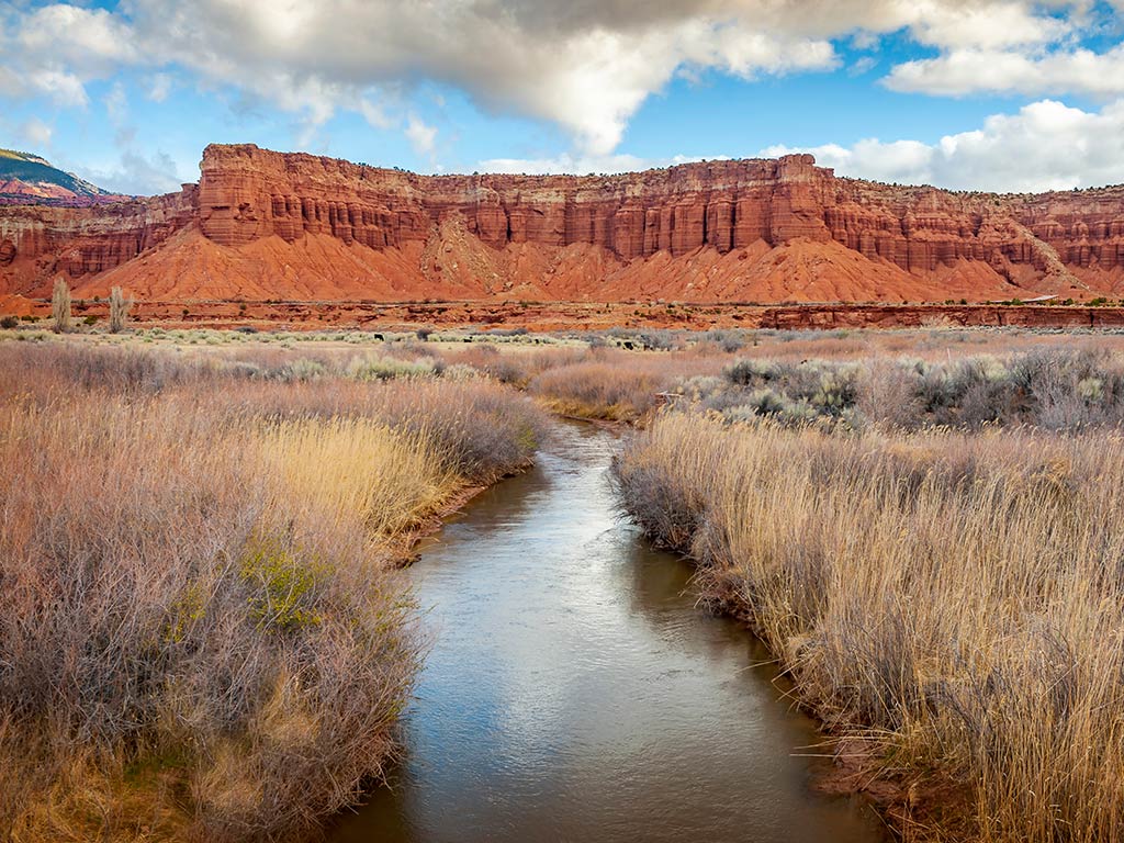 A narrow stream in Utah flows through dry grassland toward a red rock mesa under a partly cloudy sky.