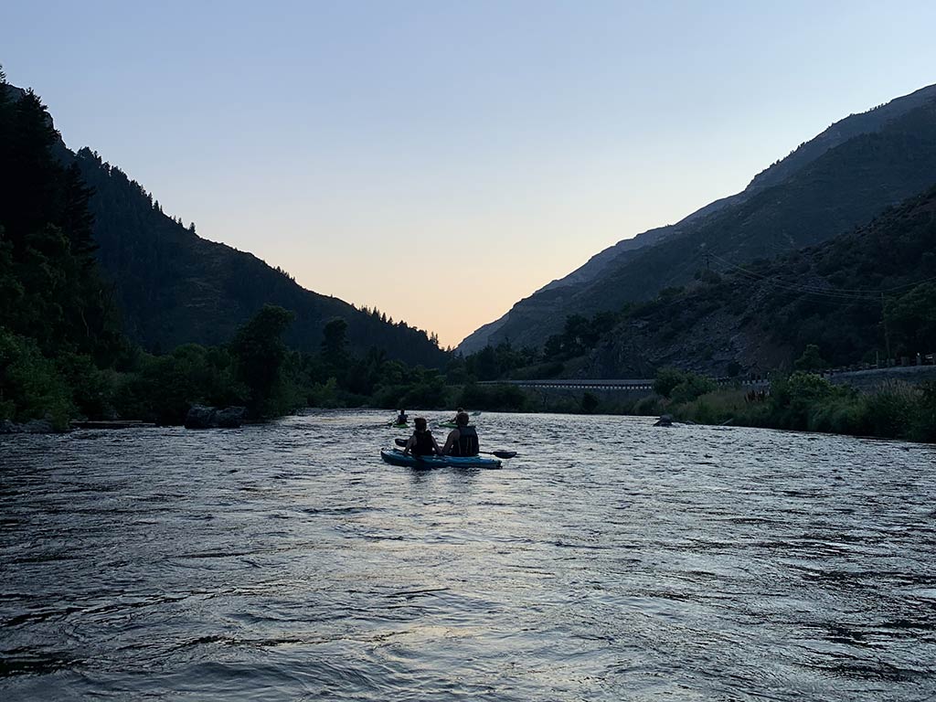A view towards a silhouette of two people in an inflatable raft floats down a river surrounded by forested hills at dusk.