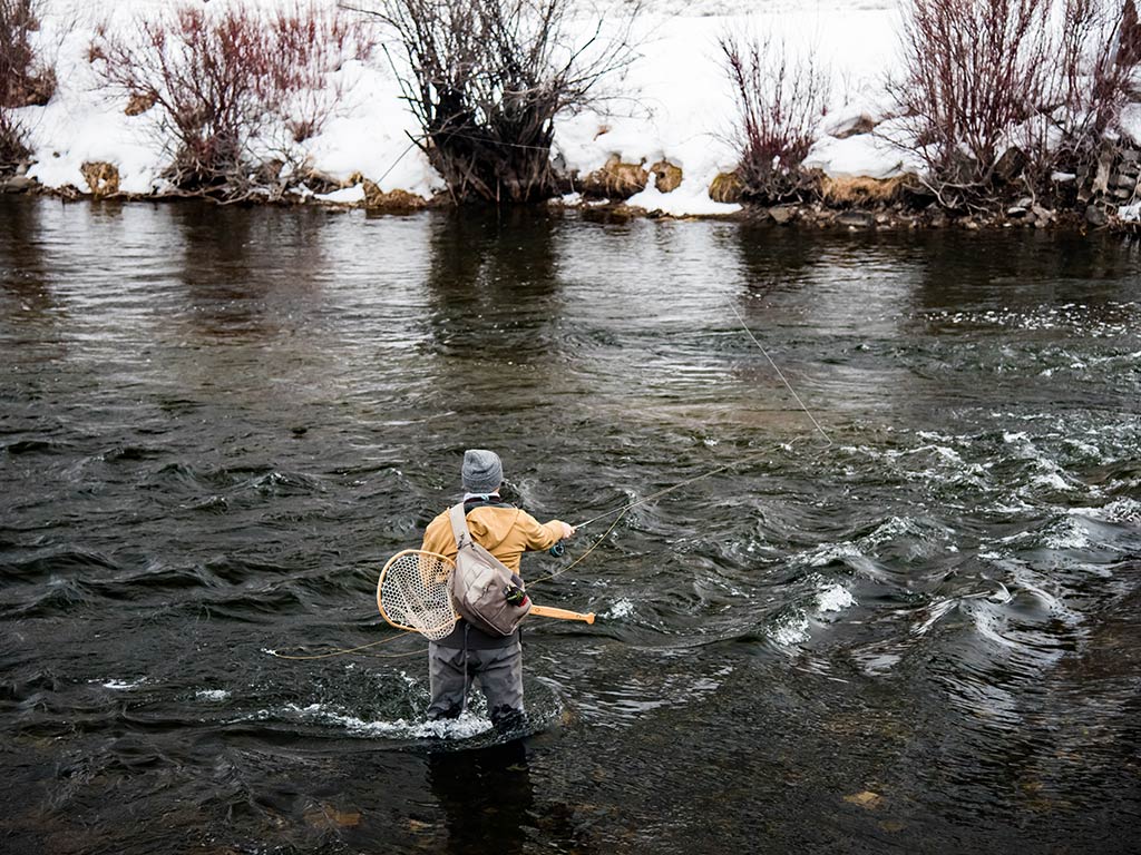 A view from behind of a person enjoying Trout fishing in Utah, casting a line into a river bordered by snow-covered banks and leafless shrubs.