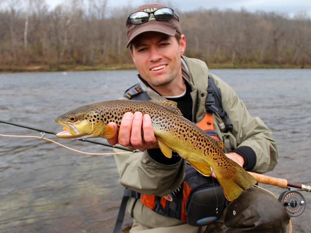 A person wearing outdoor gear holds a Brown Trout by a river in Utah, proudly displaying their catch on a cloudy day.