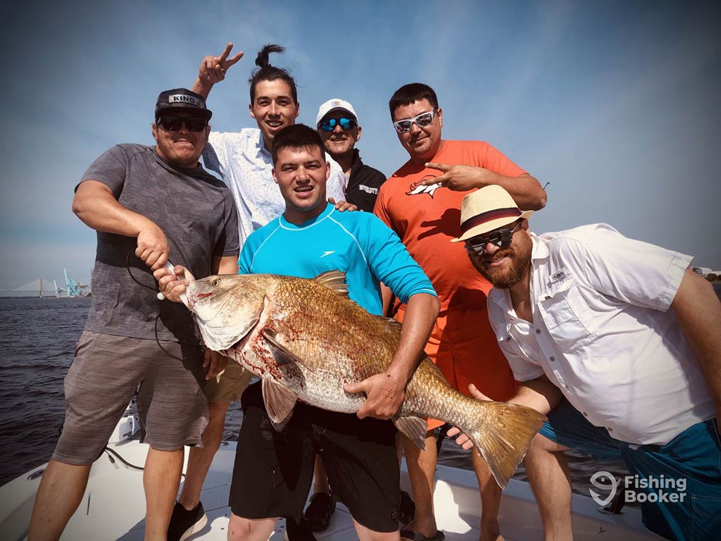 Six men are on a boat, posing for a photo, as one is holding a large Redfish. The group’s smiling at the camera on a sunny day near the water.