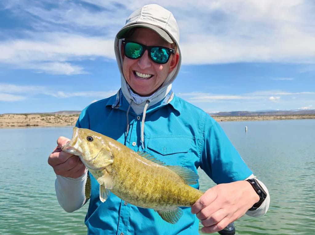 A person wearing sunglasses and a sun shirt holds a Bass on a boat on a lake in Utah, with blue skies in the background.