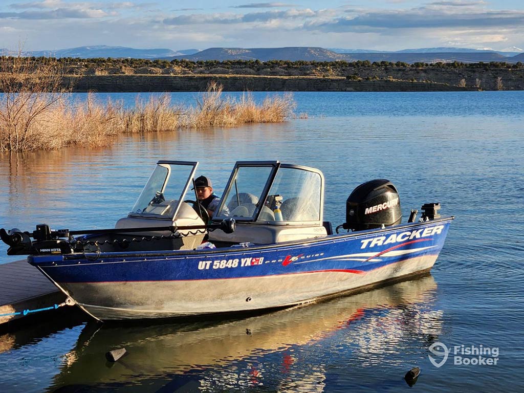 A person sitting in a blue Tracker fishing boat with a Mercury motor, docked on a calm lake in Utah. The scene is set against mountains and dry brush in the background.