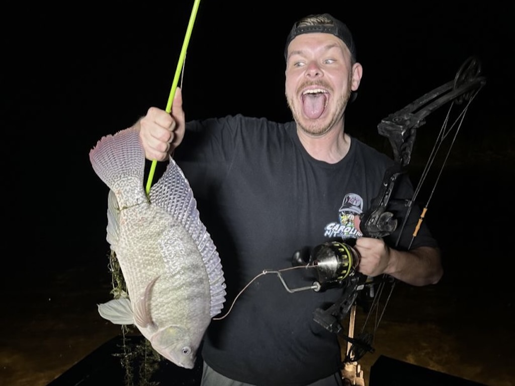 A man stands outdoors at night, smiling widely as he holds a large Tilapia on a line in one hand and a fishing bow in the other.