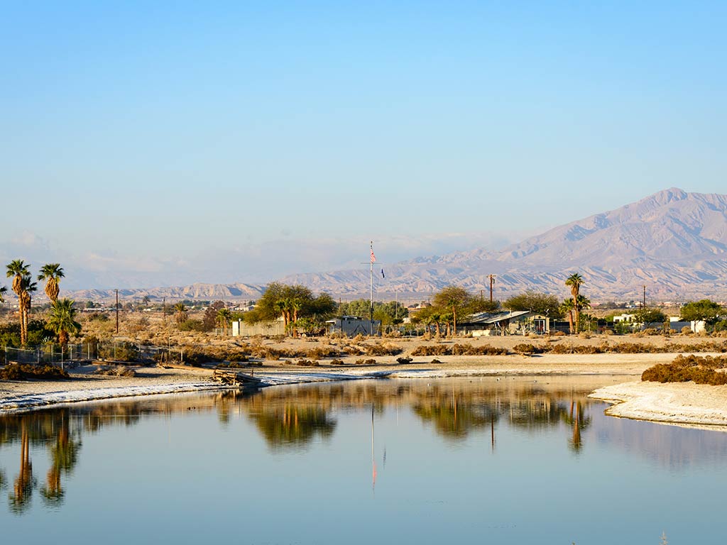 A calm body of water reflects nearby palm trees, desert vegetation, and distant mountains under a clear blue sky.