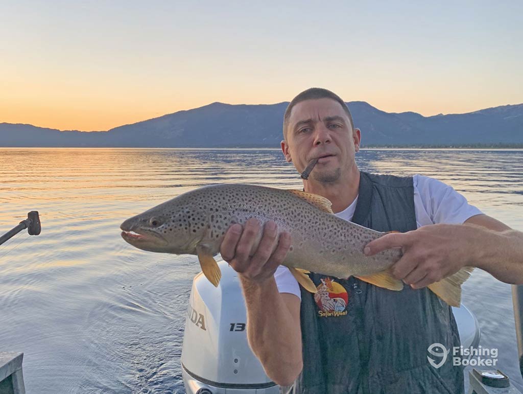 A man proudly holds a large California Trout on a boat at sunset, with majestic mountains and shimmering water in the background.