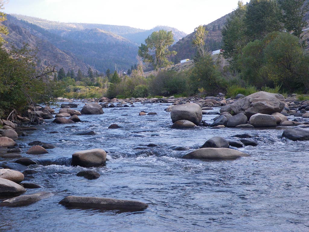 A rocky river meanders through a scenic valley, with trees lining the banks and hills in the background.
