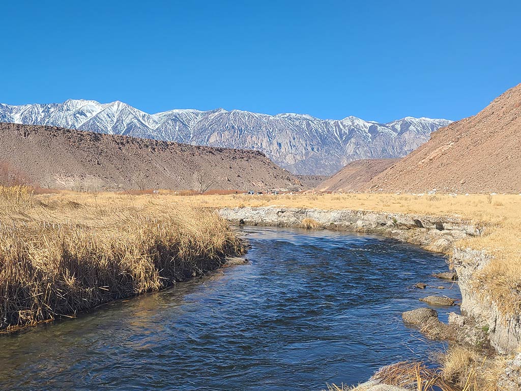 A view of the calm Owens River looking towards some snow-capped mountains, as it winds through a grassy area, framed beneath a clear blue sky.