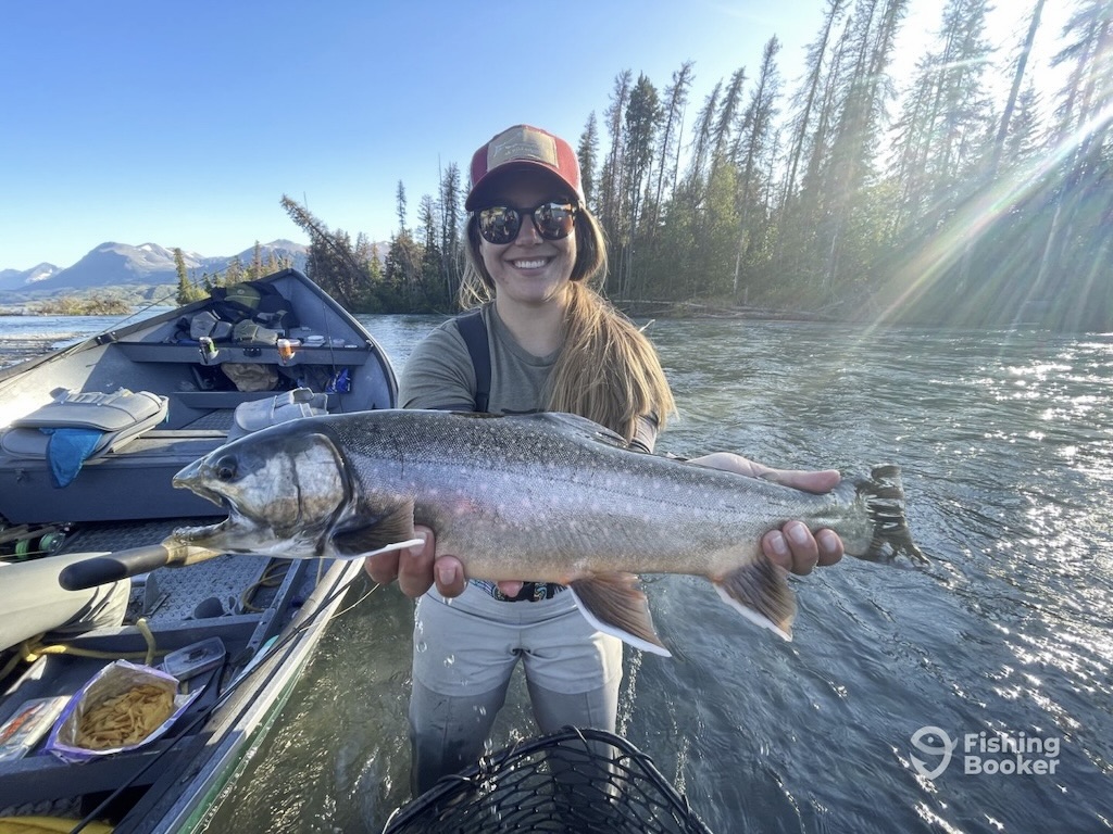 A woman is standing in a river, holding a large Char with both hands and smiling at the camera. Behind them, you’ll see a boat with fishing gear. 