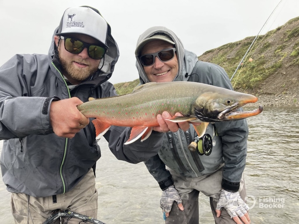 Two people in outdoor gear stand by a riverbank, smiling at the camera as they hold a large Char together. One's wearing sunglasses and a cap, while the other has glasses and a hooded jacket.