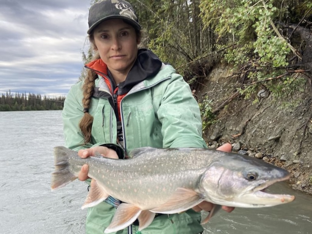 A woman in outdoor clothing is holding a large Char near a riverbank, with trees in the background. 