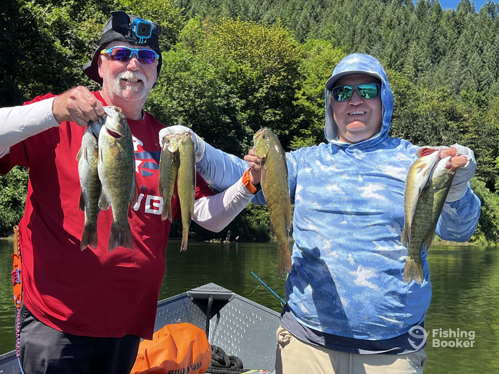 Two people are standing on a boat, proudly holding their catch from a day of Bass fishing in Oregon. They're dressed in sun-protective clothing, hats, and sunglasses, with scenic trees and water behind them.