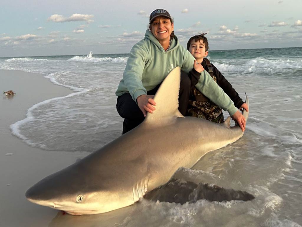 A mother and a child are kneeling on a sandy beach, posing with a large Shark they caught while shore fishing