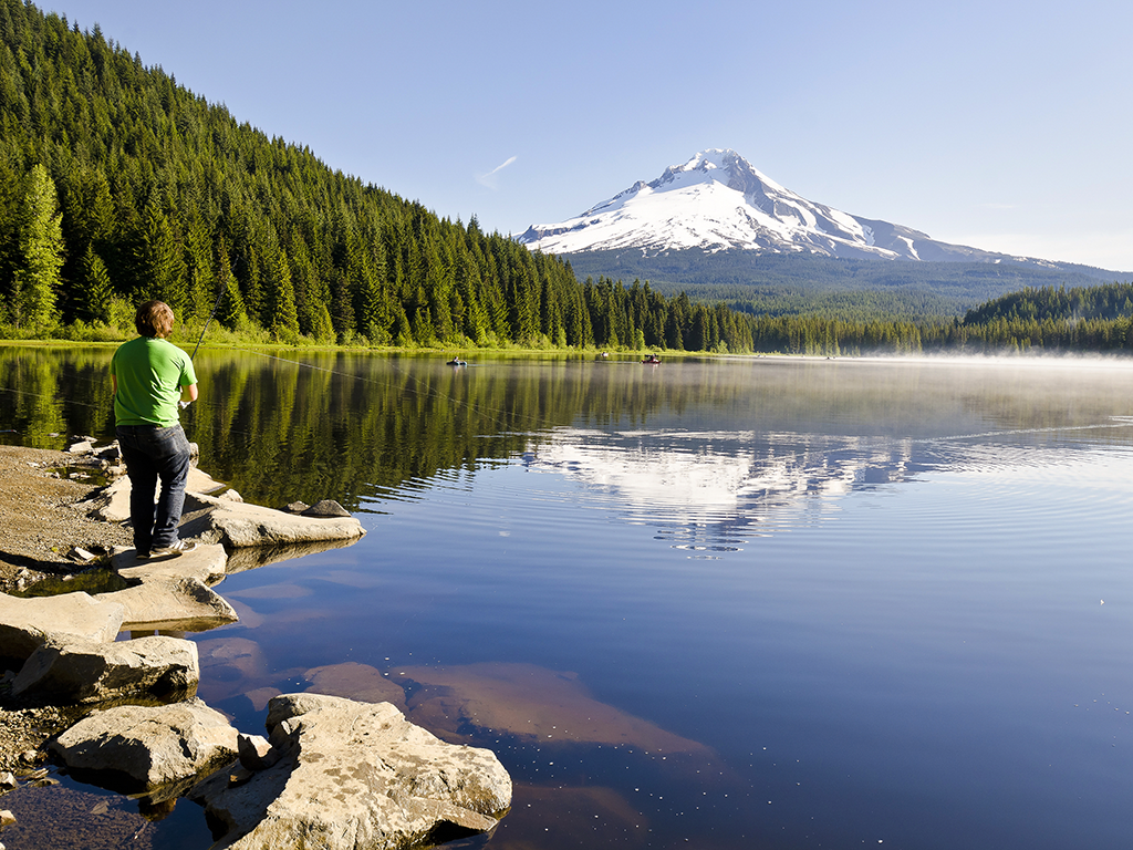 A person standing on rocks at the edge of a calm lake, Bass fishing in Oregon. A snow-capped mountain and dense pine forest are reflected in the water.
