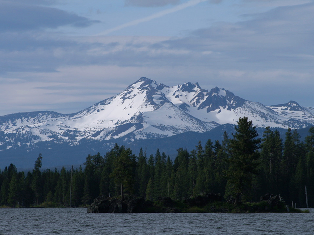 A snow-covered mountain with visible peaks rises behind a dense forest and a lake, under a cloudy sky.