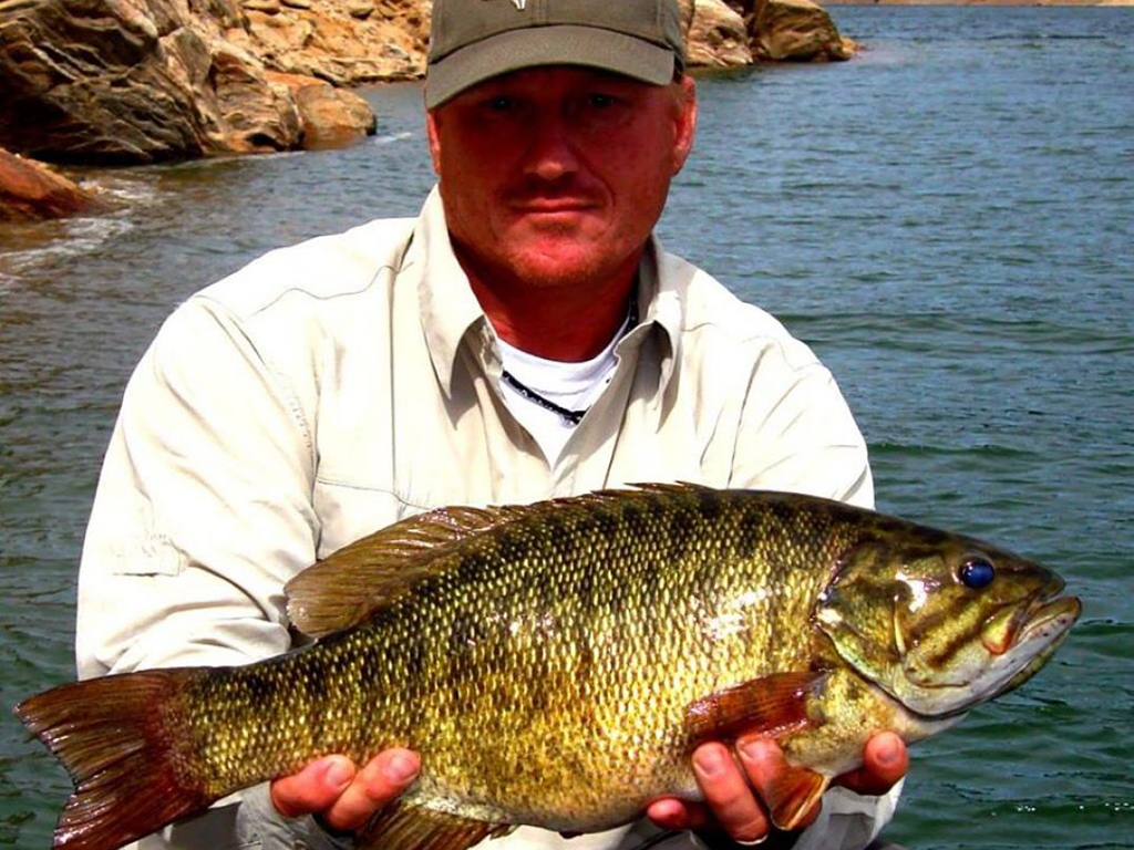 A man wearing a cap and light-colored shirt shows off a Smallmouth Bass fish he caught in Oregon near a rocky shoreline.