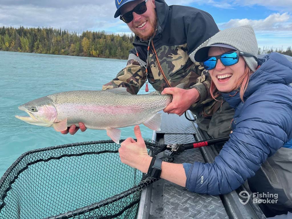 Two people on a boat are holding a large Rainbow Trout over a net above blue water. They’re both wearing sunglasses and outdoor clothing, standing in front of trees in the background.