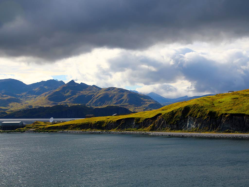 A coastline stretches along green hills under a cloudy sky. In the foreground, water gently laps against the shore, while majestic mountains rise in the background.