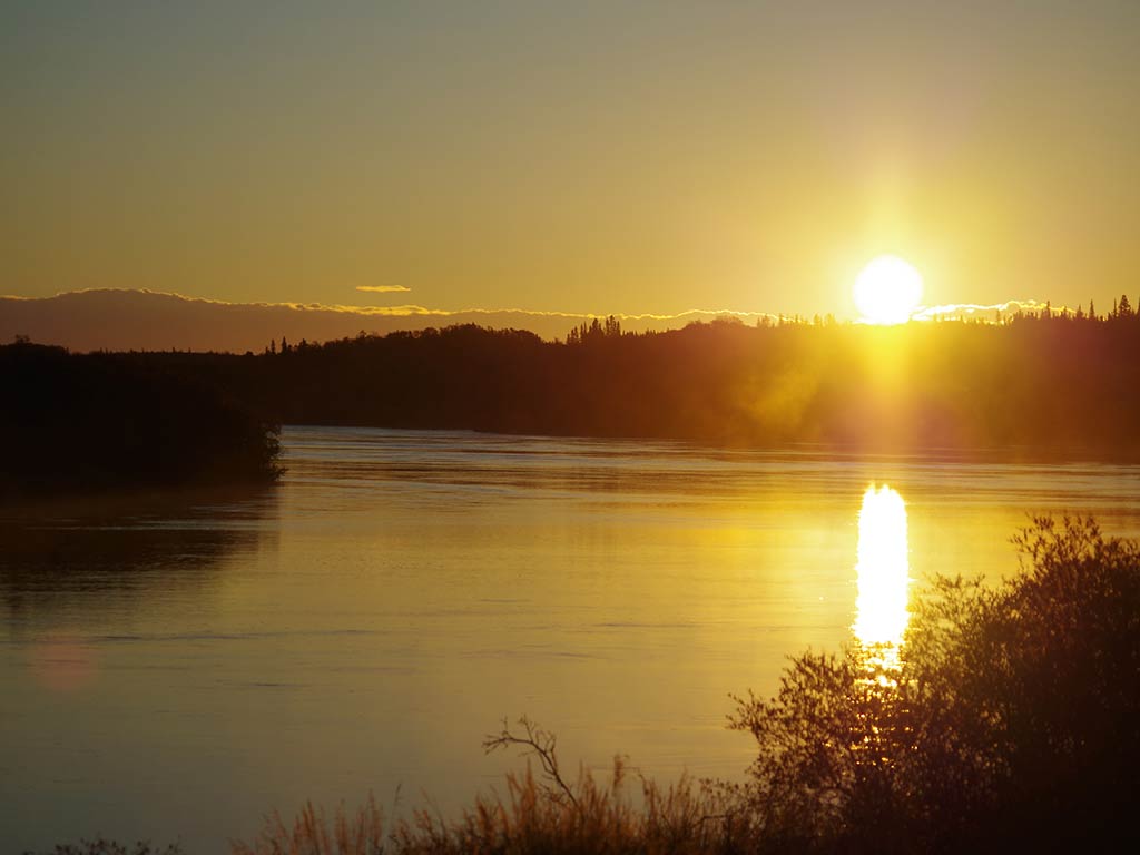 The sun sets over the calm Kvichak River, casting a bright reflection on the water, with silhouetted trees along the horizon.